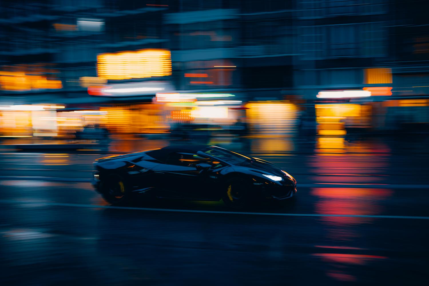 Coche deportivo negro circulando a alta velocidad por una ciudad iluminada con neones durante la noche.
