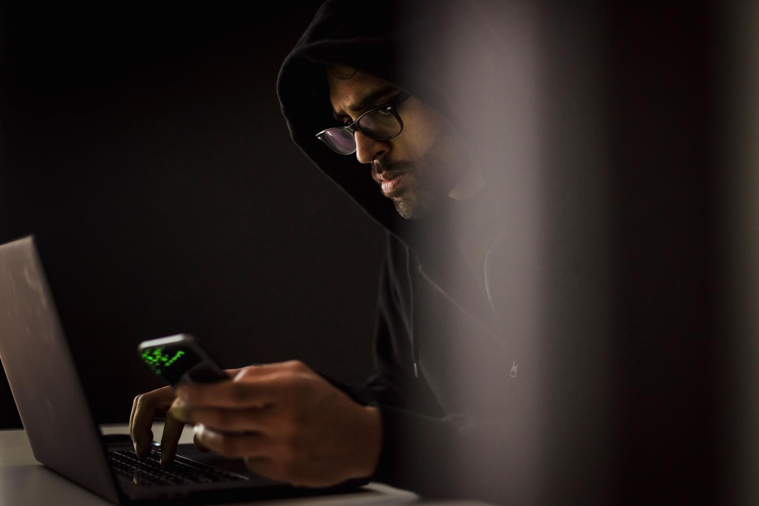Ciberdelincuente con capucha en una habitación oscura utilizando un portátil y un smartphone para realizar ataques informáticos.
