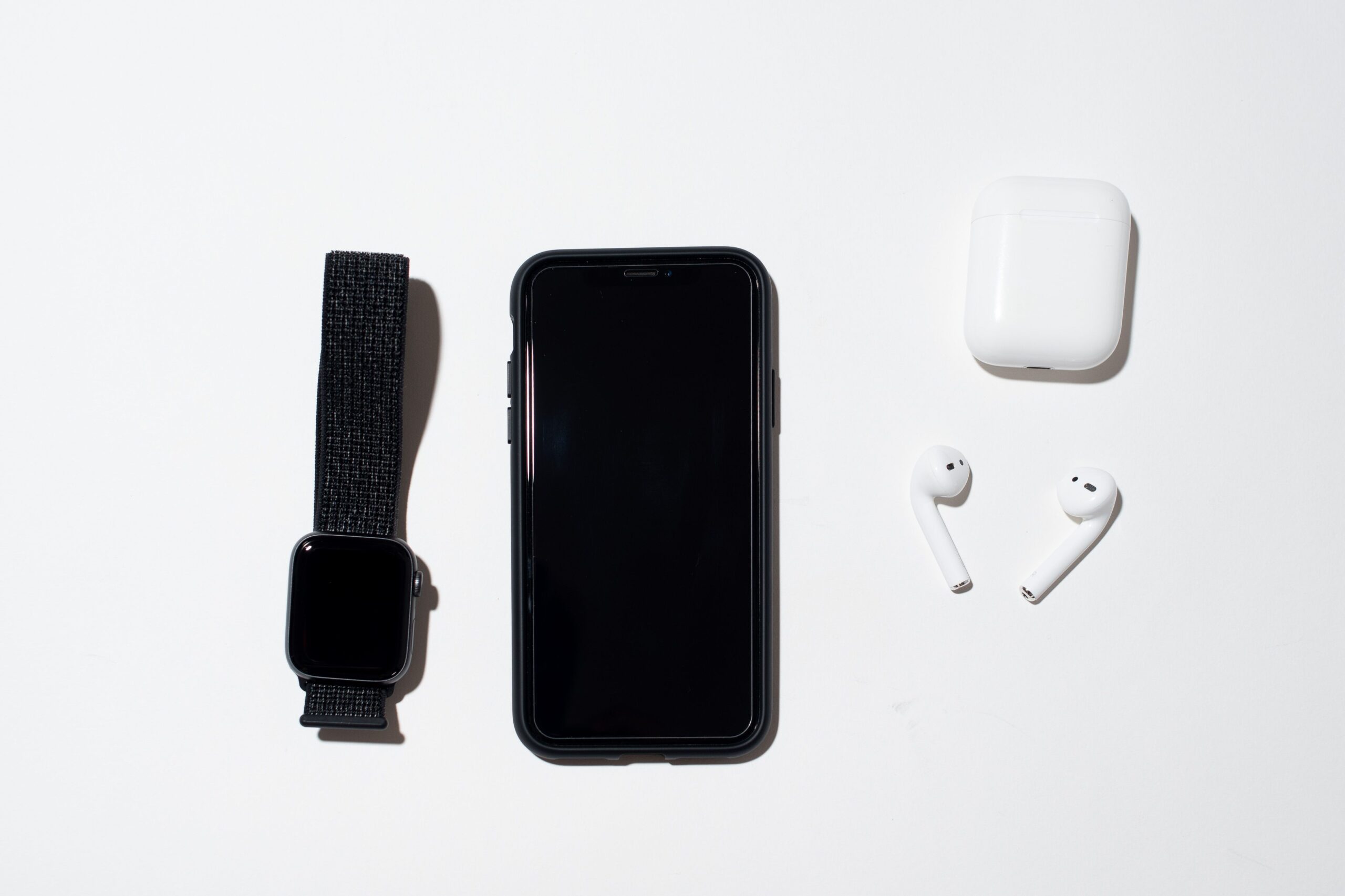 Vista cenital de un iPhone, un Apple Watch y auriculares inalámbricos blancos organizados sobre un fondo blanco limpio.