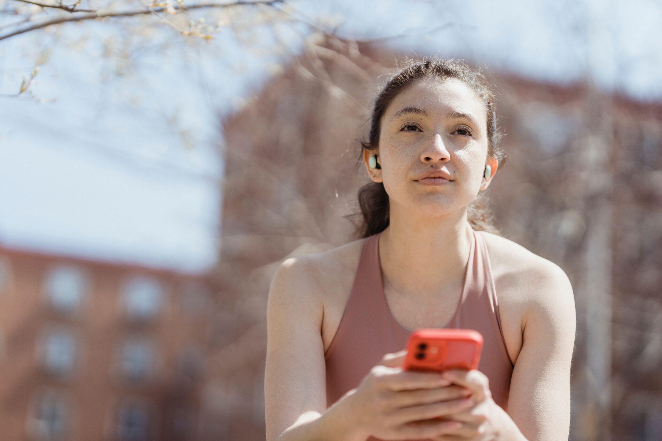 Mujer joven utilizando auriculares inalámbricos y un smartphone en un entorno exterior soleado, capturando un estilo de vida tecnológico y natural.