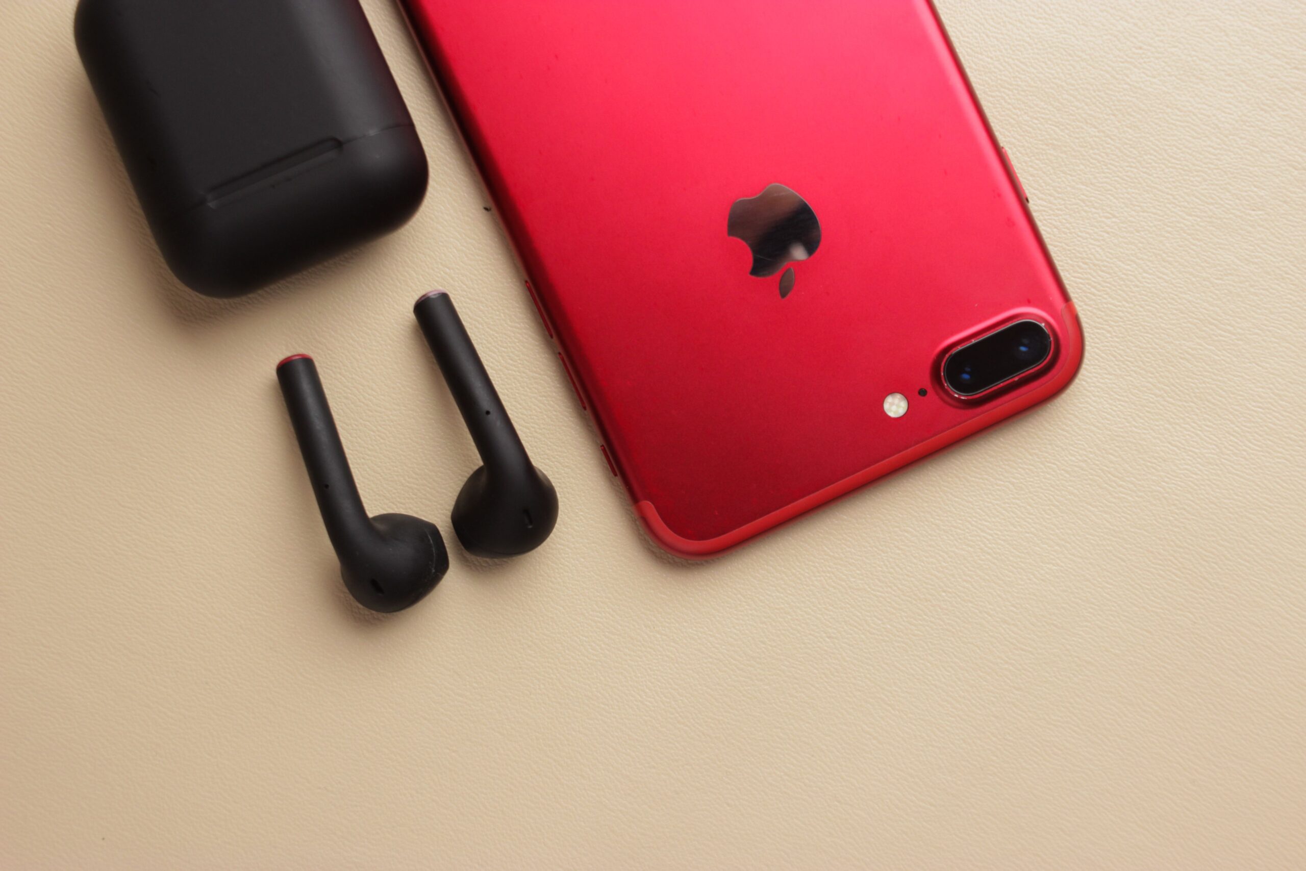 Primer plano de un iPhone rojo junto a auriculares inalámbricos negros y su estuche de carga sobre una superficie neutra.