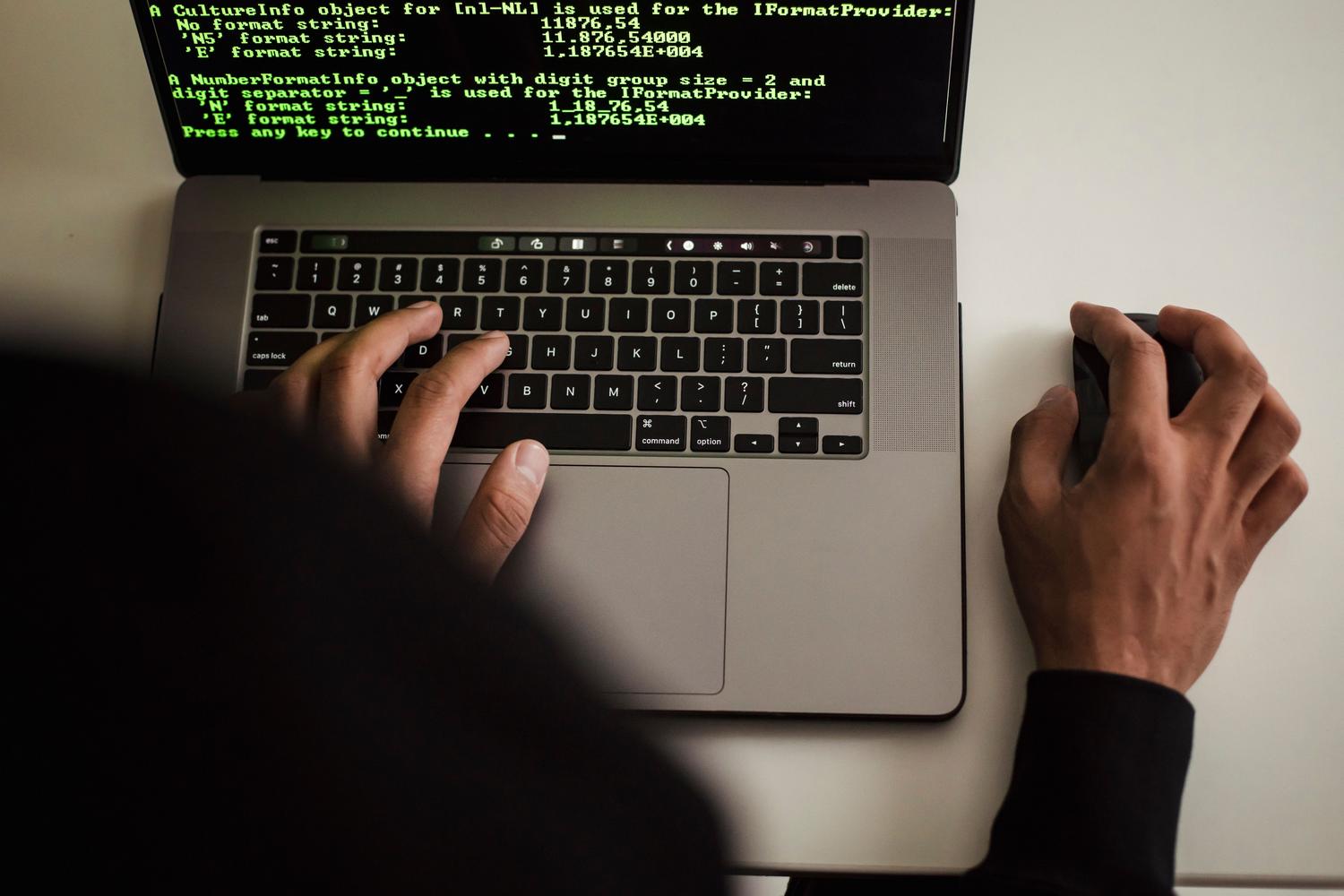 Persona encapuchada trabajando en un ordenador portátil con código en pantalla, representando a un ciberdelincuente.