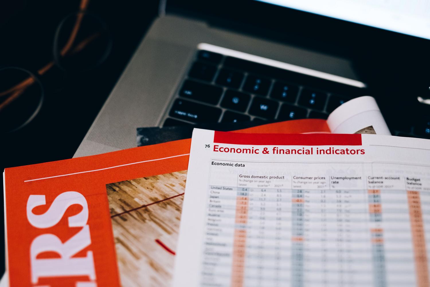 A close-up, high-angle shot of a professional desk setup. In the foreground, a printed report displays a table of 'Economic & financial indicators' with columns for GDP and unemployment rates. In the blurred background, a laptop keyboard and eyeglasses suggest an office environment focused on data analysis.