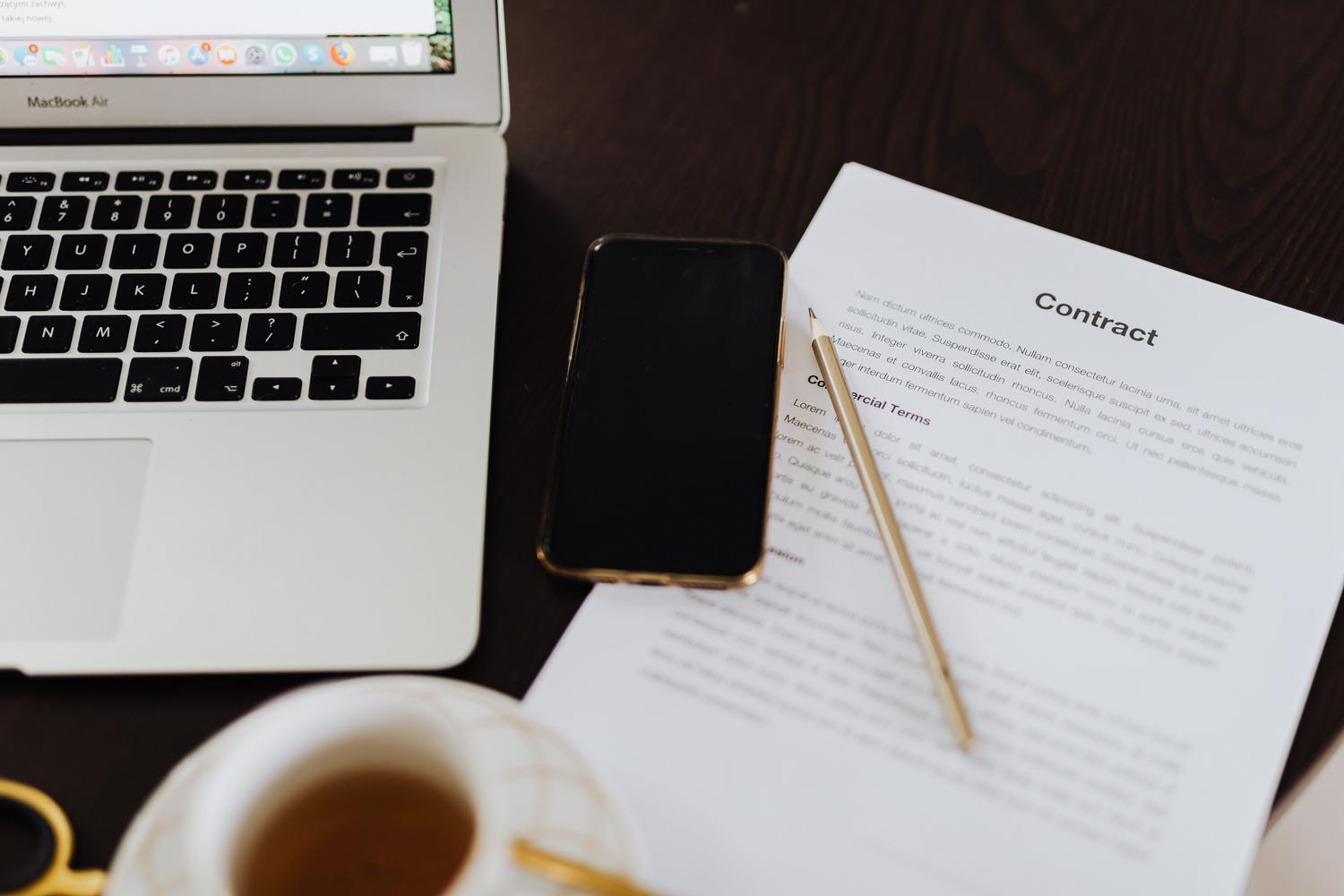 Espacio de trabajo moderno con un contrato, ordenador portátil y smartphone, representando la sincronización de archivos en la nube y multidispositivo.