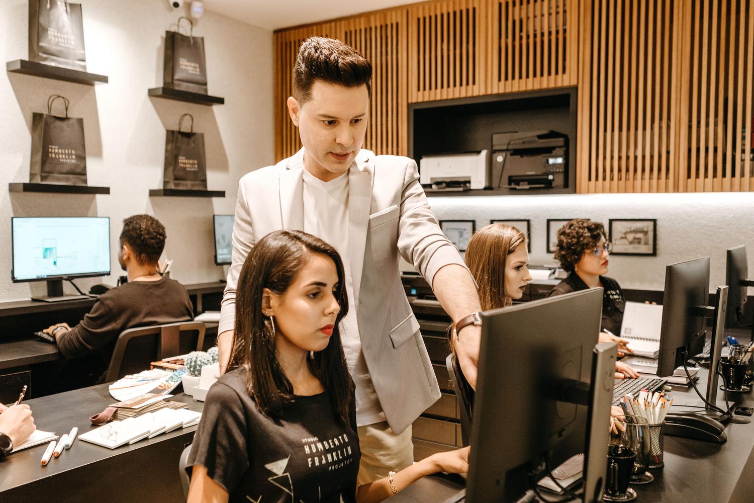 Un mentor en una oficina moderna enseñando a una empleada frente al monitor, representando el microaprendizaje y la formación 'justo a tiempo'.