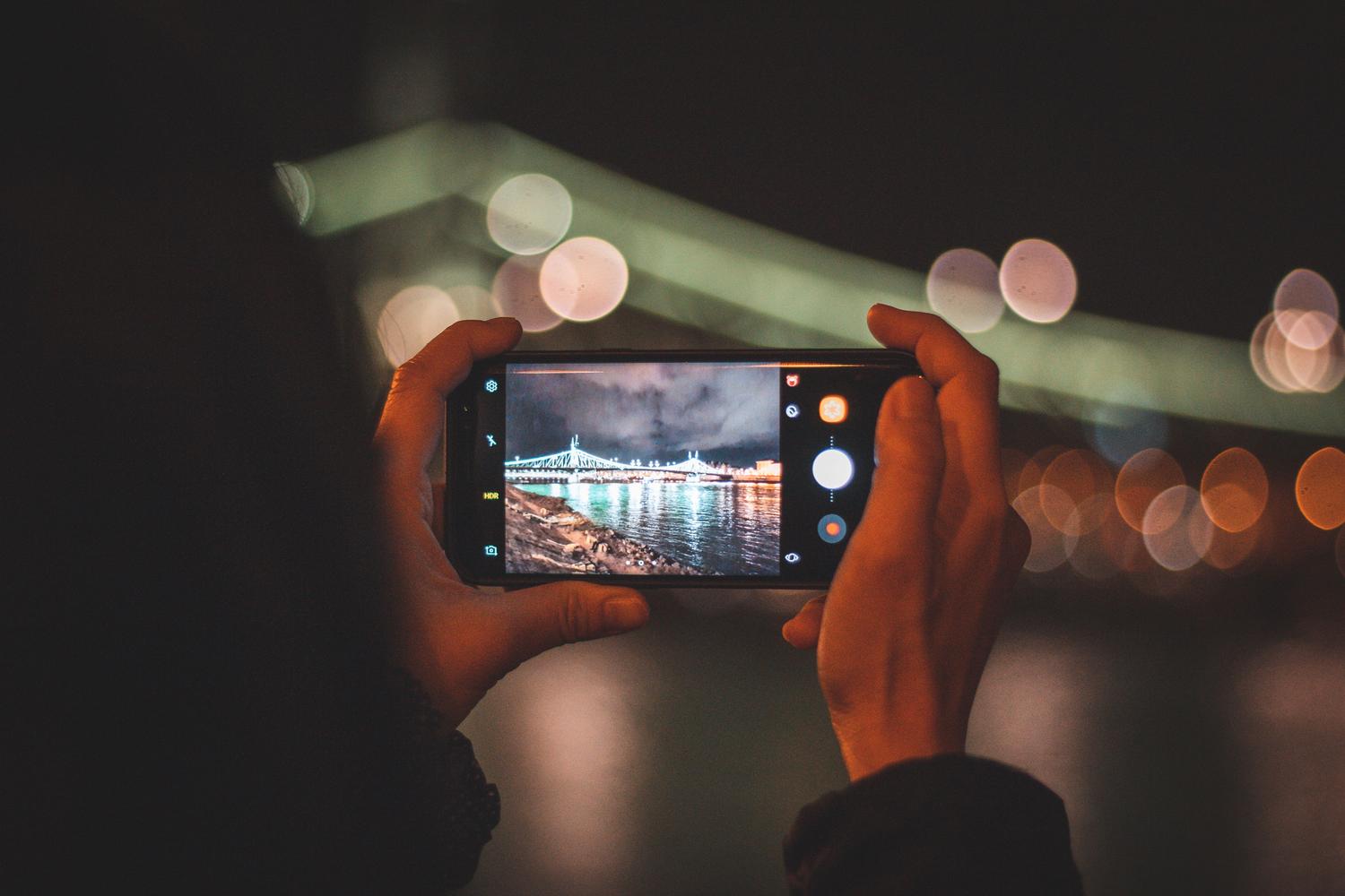 Persona fotografiando un puente iluminado de noche con un smartphone, destacando el efecto bokeh del fondo.