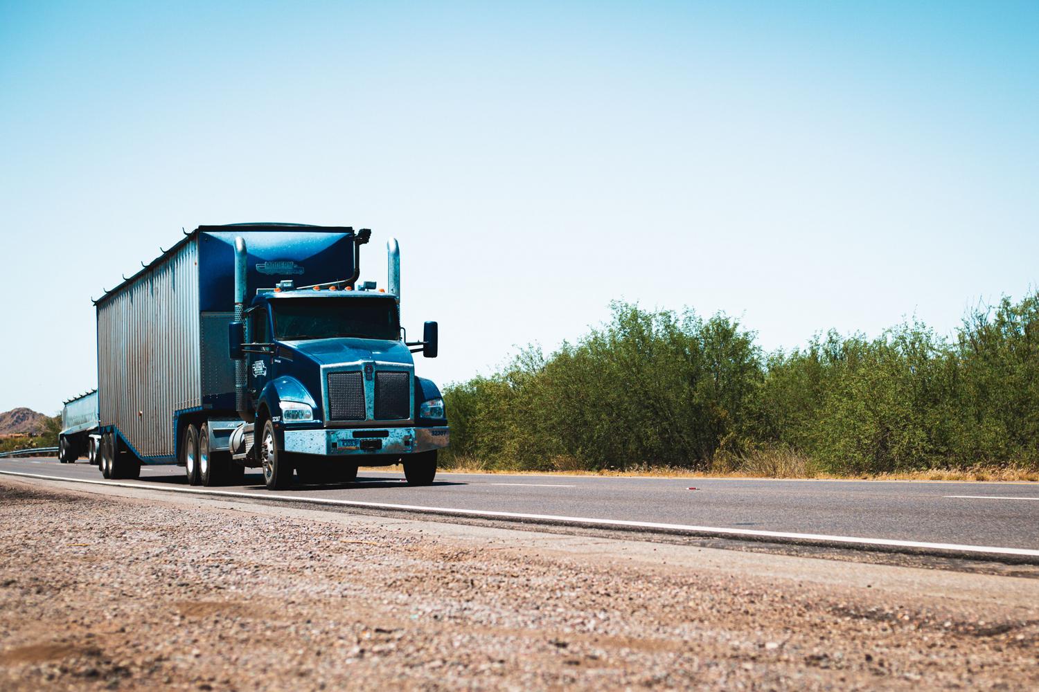 Camión semi remolque azul circulando por una carretera rural de Estados Unidos, ilustrando la conducción inmersiva en rutas secundarias Camión semi remolque azul circulando por una carretera rural de Estados Unidos, ilustrando la conducción inmersiva en rutas secundarias