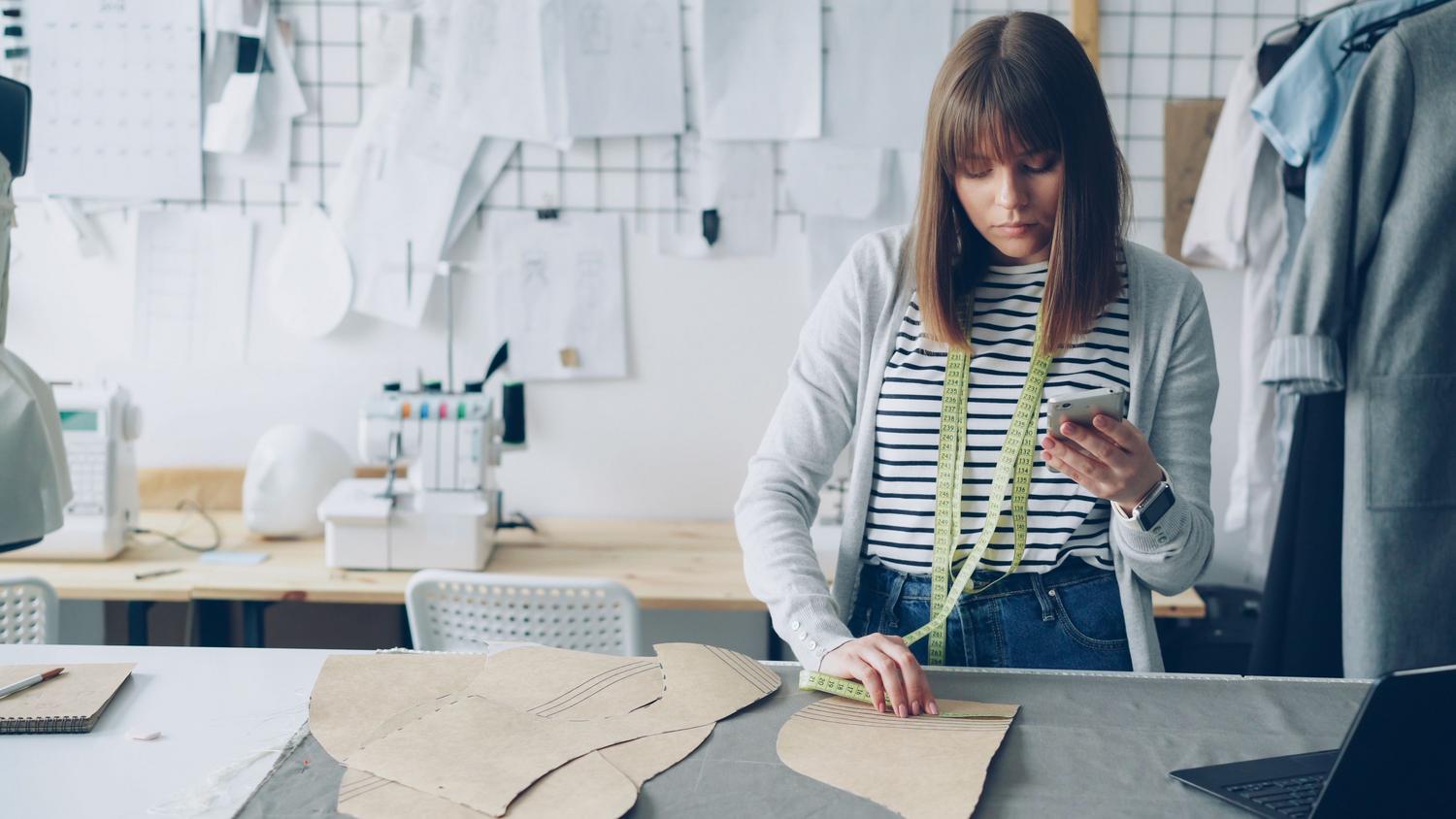 Diseñadora de moda integrando el uso de un smartphone con herramientas de costura tradicionales en su estudio.