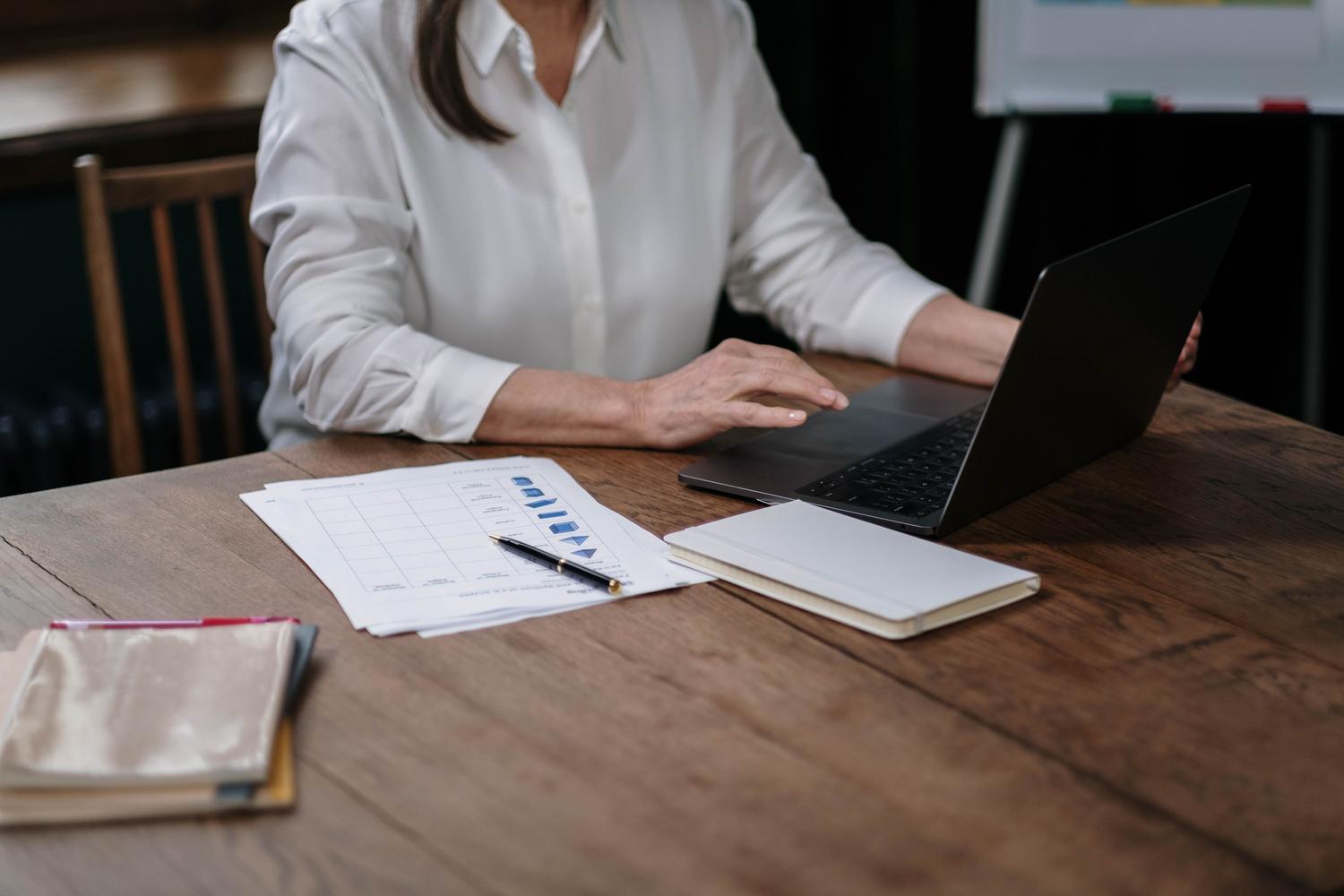Mujer de negocios analizando datos en un portátil con documentos impresos en formato de tabla.