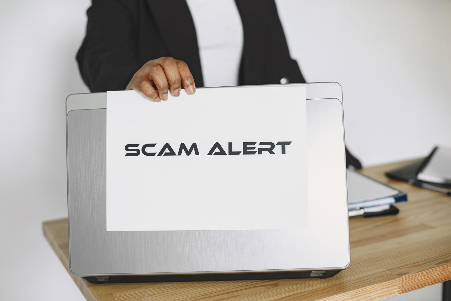 A person in professional attire holding a paper sign that says "SCAM ALERT" over the back of a laptop screen, illustrating cybersecurity and phishing detection.