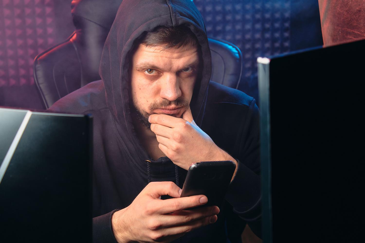 A man in a black hoodie using a smartphone in a dark, digital environment, representing the anonymous nature of cybercriminals sending phishing attacks.