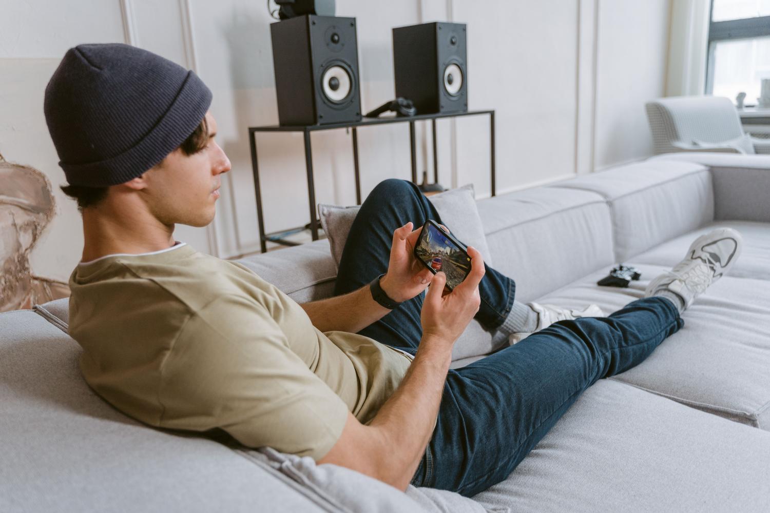 Hombre relajado en un sofá jugando con su móvil, ejemplificando la portabilidad de llevar la consola PS3 en el bolsillo.
