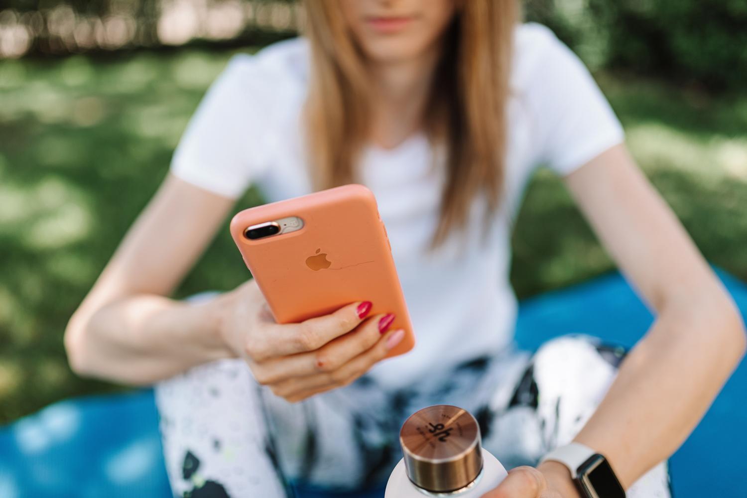 Mujer joven utilizando su iPhone en un parque soleado, ilustrando la personalización del dispositivo en entornos sociales y viajes.